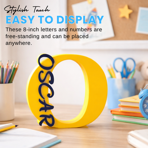 A yellow freestanding decorative letter 'O' with the name 'OSCAR' in dark blue script, displayed on a desk with school supplies in a classroom-like setting. Text at the top reads 'Stylish Touch EASY TO DISPLAY' and describes the letters/numbers as free-standing and placeable.