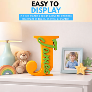A freestanding decorative wooden letter J in orange with green script lettering spelling 'James', displayed on a white dresser in a styled room with a lamp, teddy bear, picture frame, and star decor. Text at the top reads 'EASY TO DISPLAY'.